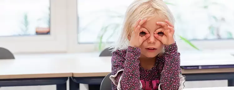 Blond meisje zit aan haar tafel en maakt een bril met haar vigers