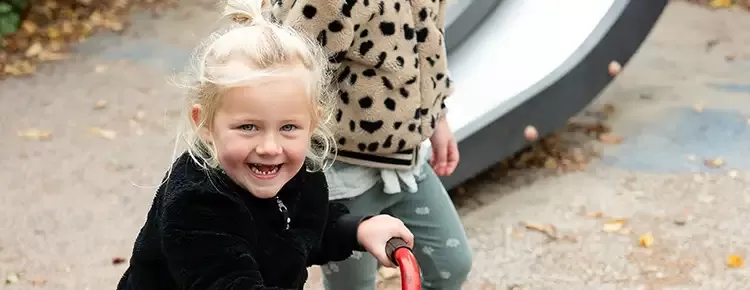 meisje kijkt lachend de camera in terwijl zij op een driewielfiets op het schoolplein zit