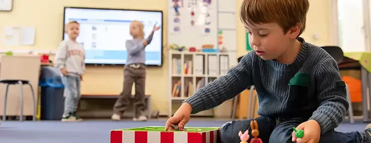 Jongen van een jaar of 5 speelt met figuurtjes van Kikker terwijl 2 klasgenootjes op de achtergrond spelen