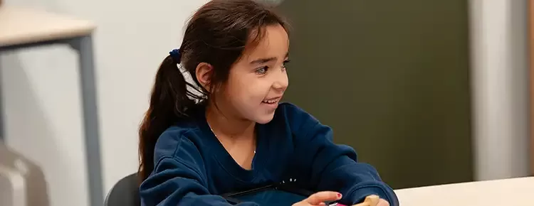 Meisje van een jaar of 8 met bruin haar en een blauwe trui zit aan haar tafel in de klas